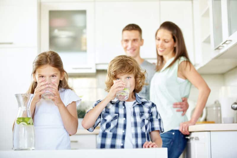 a mother, father, daughter, and son standing in a kitchen drinking water from glass cups