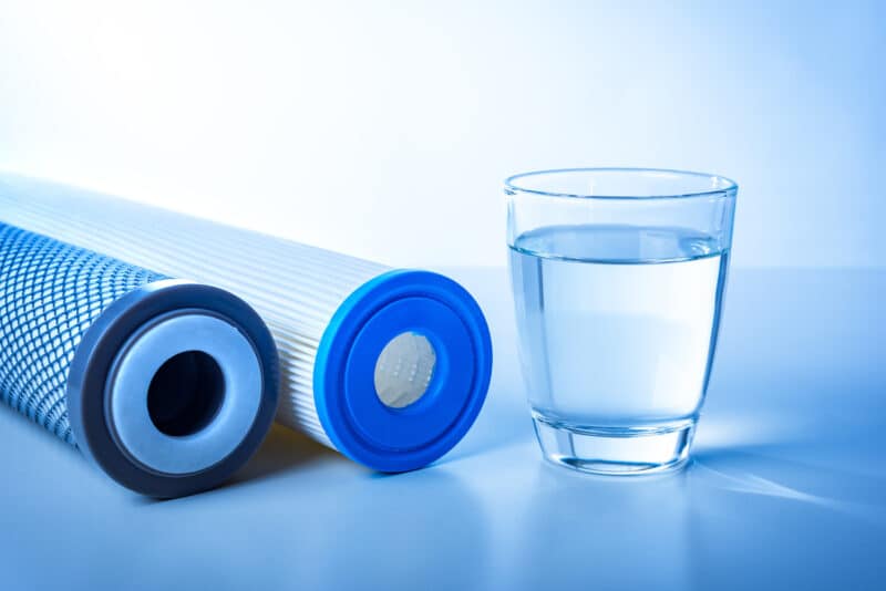 two water filtration cartridge laying on a blue table beside a short glass cup full of water