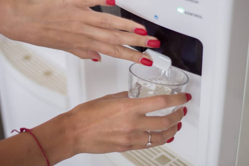 woman with red nail polish filling up a small glass cup with water from a cooler dispenser