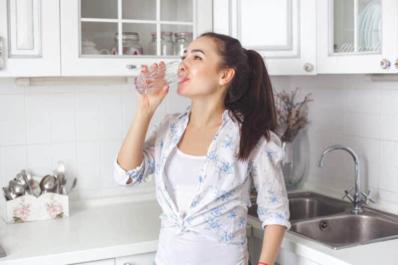 Young healthy woman drinking filtered water from a glass cup in her kitchen