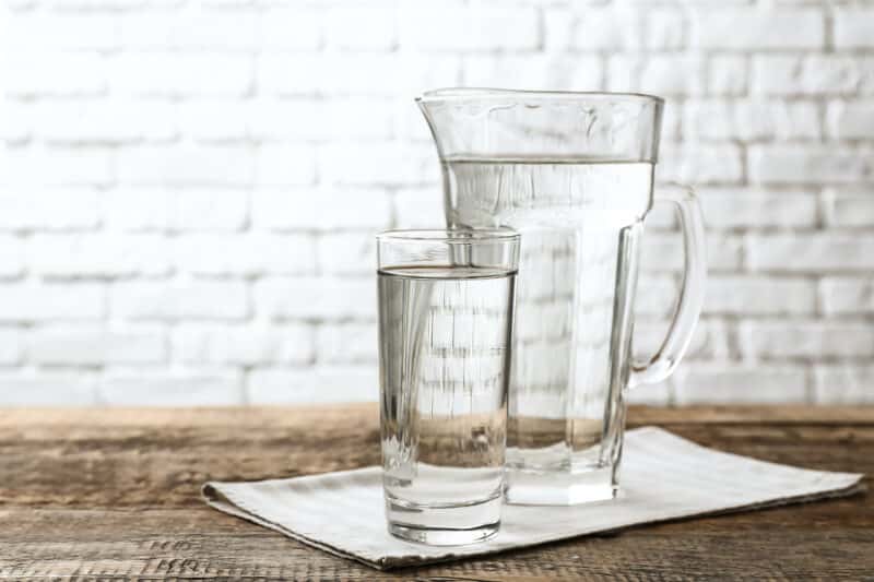 a glass cup and pitcher full of water, sitting on a wooden dining table in front of a white brick wall