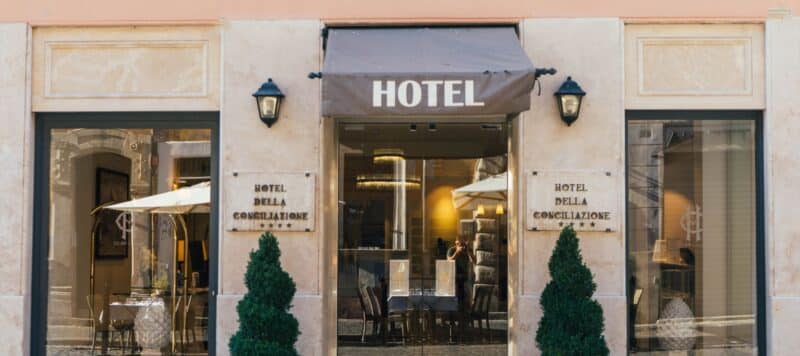 Facade of Hotel Della Conciliazione, featuring a central entrance with a brown awning labeled 'HOTEL.' The entrance is flanked by two symmetrical windows, potted greenery, and elegant outdoor seating. The exterior showcases classic architectural details and warm lighting, inviting guests inside.