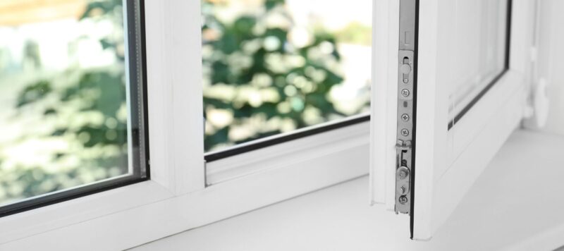 A close-up of an open white-framed tilt-and-turn window with visible metal locking mechanism and blurred greenery outside
